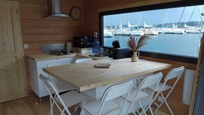 A compact kitchen with a wooden table and white chairs, a stove, a microwave, and a clock on the wall. A window offers a view of boats at a marina.