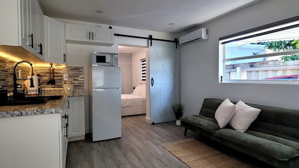 Cocina compacta con armarios blancos, microondas y refrigerador. Una puerta corrediza da acceso a un dormitorio con cama y una planta en maceta. Un sofá verde está apoyado en la pared, y hay una ventana con vistas al exterior.