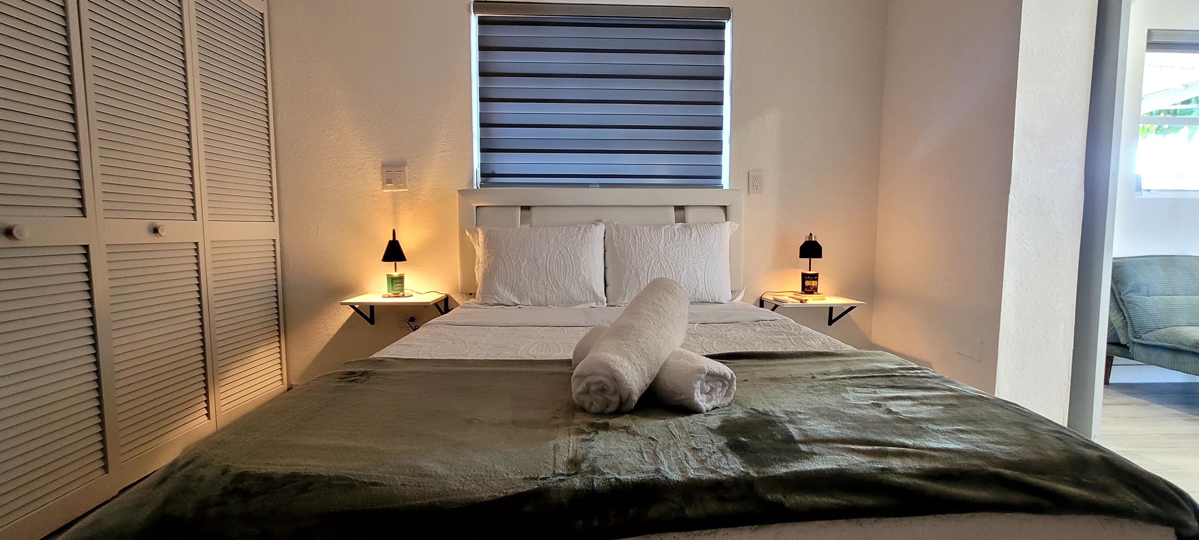 Une chambre à coucher avec un lit, deux lampes de chevet, une fenêtre avec des stores et une vue sur une chaise à l’extérieur.
