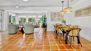 Un amplio salón con una mesa de comedor preparada para seis personas, rodeada de sillas de mimbre. Se aprecian un ventilador de techo y lámparas colgantes sobre la mesa.
