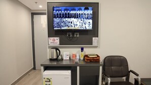 Un pequeño espacio de oficina con un televisor que muestra una multitud, una cafetera, un refrigerador y un cesto de basura.