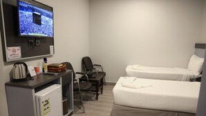 Habitación de hotel con televisión mostrando una multitud, un escritorio con hervidor, refrigerador, dos camas y una silla.