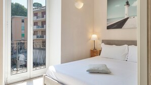 A hotel room with a bed, a nightstand with a lamp, a balcony with a view of buildings, and a painting of a lighthouse. - Casa Vico delle Isole by Wonderful Italy (Recco)
