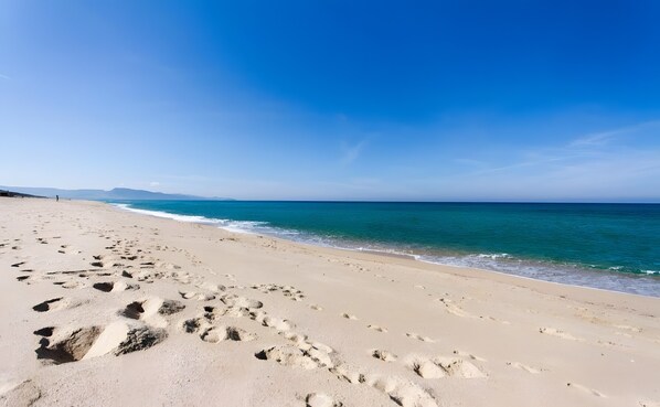 Ein Strand mit Fußabdrücken, die zum Wasser führen, ein klarer Himmel und ferne Berge.