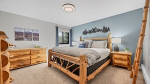 A bedroom with a wooden bed, a wooden dresser, a nightstand with a lamp, and a wall-mounted light fixture. - Condo with Ski Locker | Peak Views (Breckenridge)