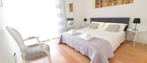 A neatly arranged bedroom with a large bed, bedside tables, a chair, and a bookshelf.