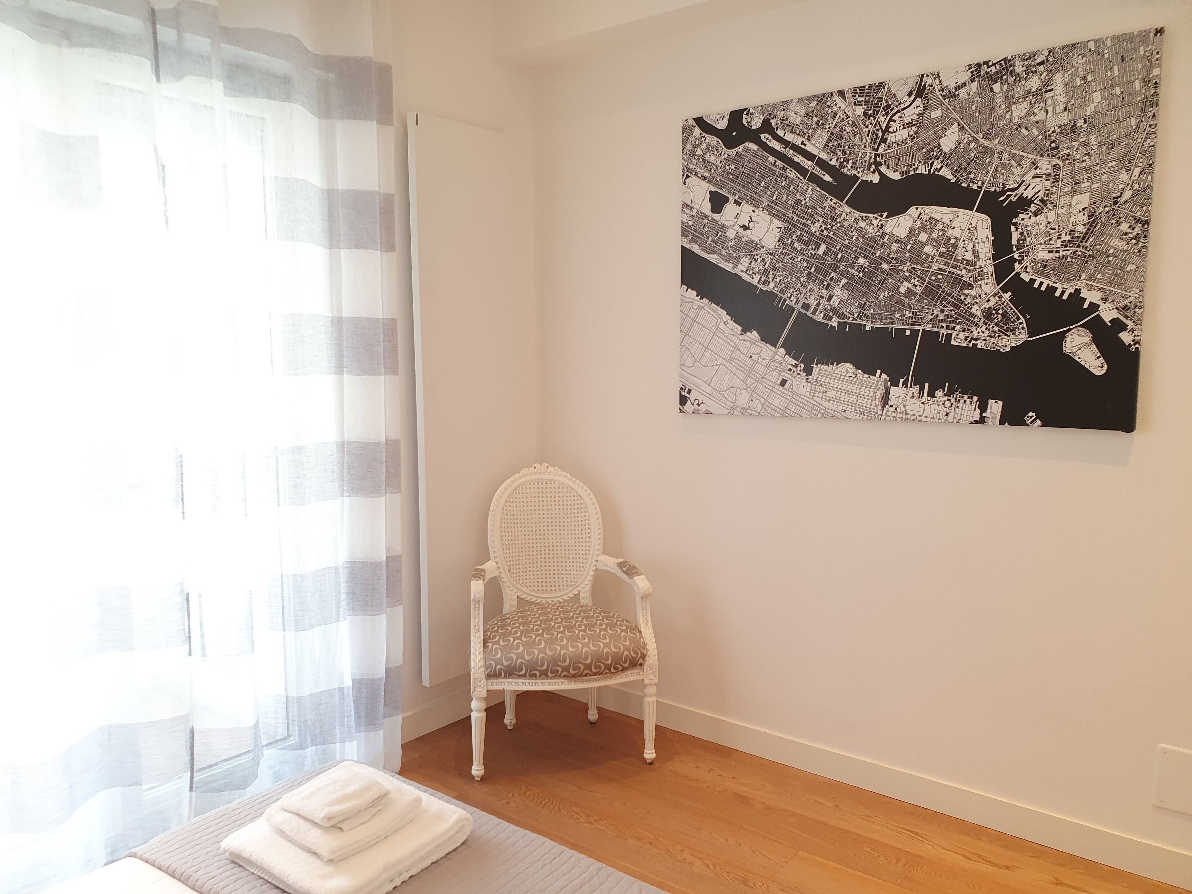 Una habitación con una gran obra de arte de un paisaje urbano en la pared, una silla blanca y una ventana con cortinas translúcidas.