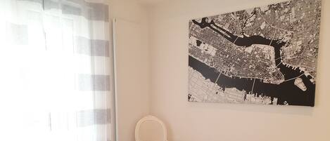 Una habitación con una gran obra de arte de un paisaje urbano en la pared, una silla blanca y una ventana con cortinas translúcidas.