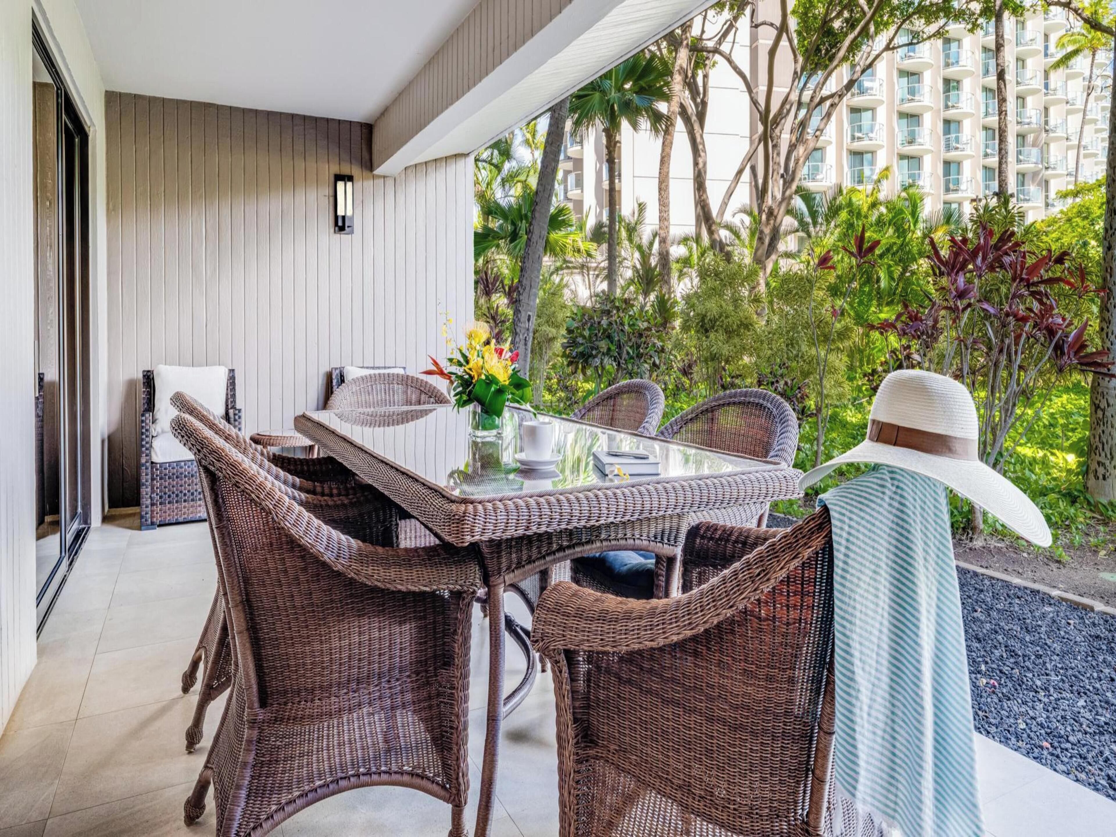 A dining area with wicker furniture and a glass table, overlooking a lush garden and a building with balconies.