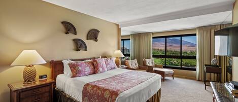 Habitación de hotel con una cama grande, dos mesitas de noche, un escritorio y una silla. Se observan ventiladores decorativos en la pared y una vista de las montañas a través de la ventana.