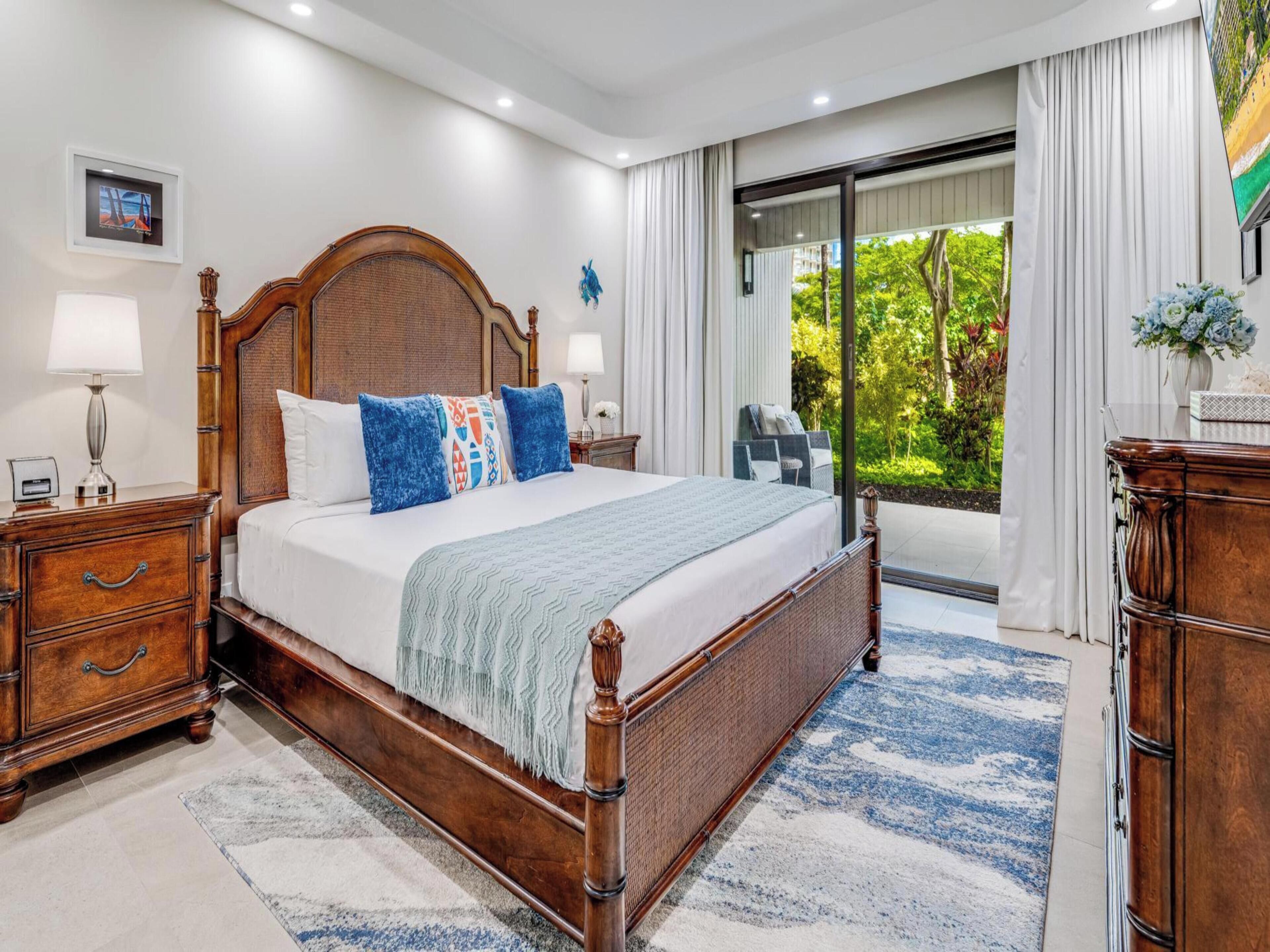 A spacious bedroom with a large bed, wooden headboard, and a view of greenery through a sliding glass door.