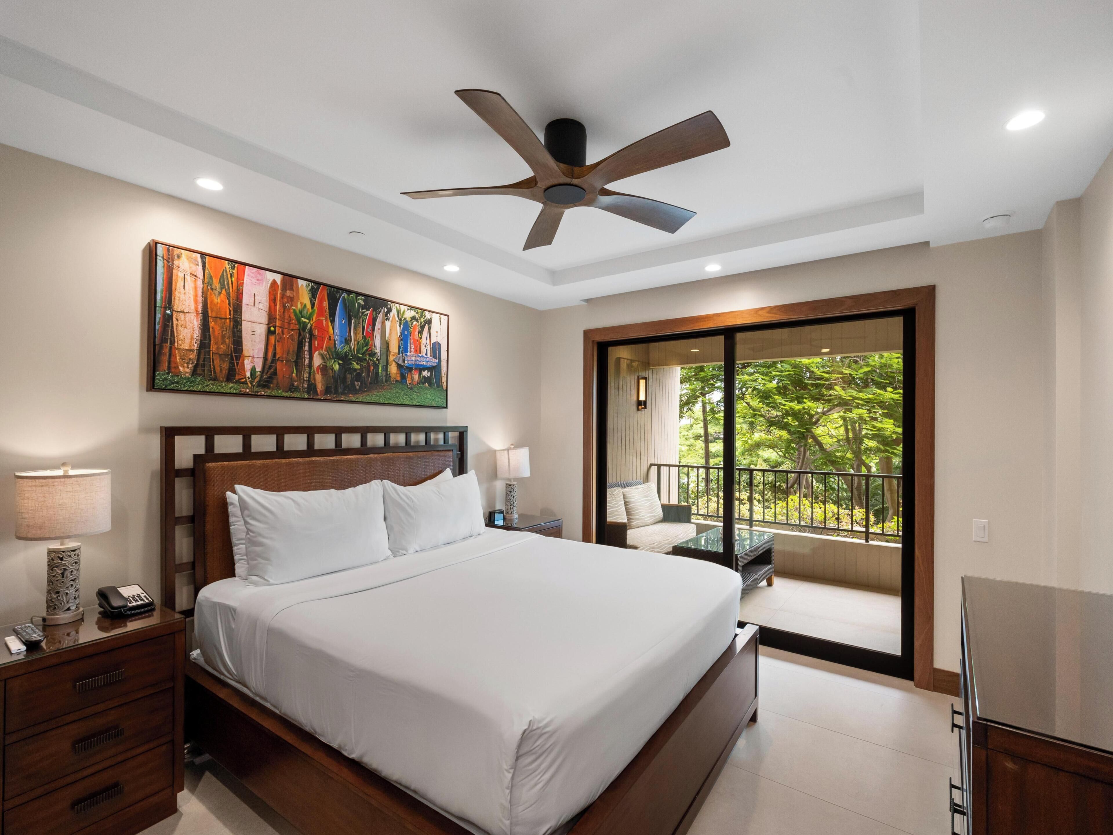 A modern hotel room with a large bed, a ceiling fan, a painting of surfboards, a balcony with a view of trees, and a TV.