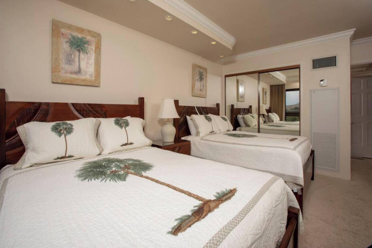 Een hotelkamer met twee bedden, elk met een palmboommotief, een nachtkastje met een lamp, een spiegel en een deur met een ventilatie rooster.