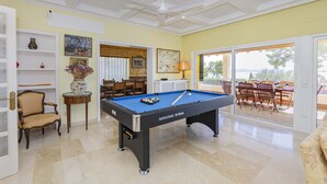Uma mesa de bilhar em um ambiente com vista para o exterior, uma área de refeições e uma estante de livros.