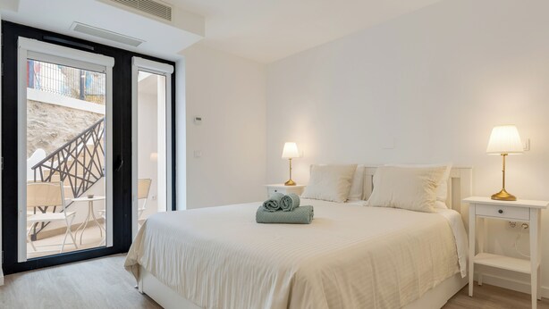 Ein Schlafzimmer mit einem Bett, zwei Nachttischen mit Lampen, einem Fenster mit Blick auf einen Balkon und einer Tür zu einem Außenbereich.