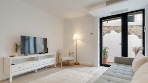Ein modernes Wohnzimmer mit Flachbildfernseher, einem weißen Sofa, einer Stehlampe und einem großen Fenster mit Blick auf ein Gebäude.