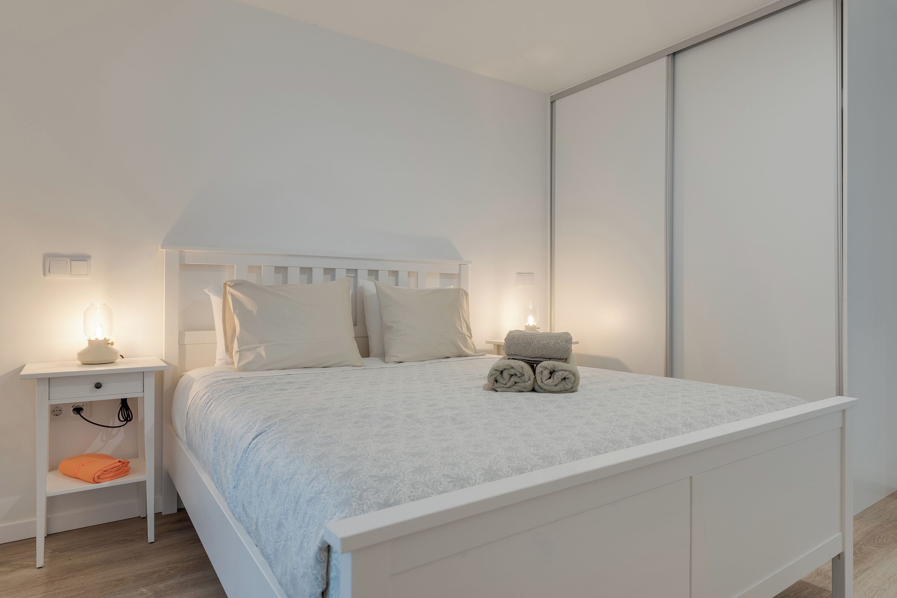 A neatly made bed with a white headboard and pillows, a bedside table with a lamp, and a rolled towel on the bed.