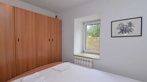 A bedroom with a large wooden wardrobe, a single bed with white linens, a radiator, a small window with a stone wall view, and a framed artwork on the wall. - Studio Veli  A in Veli Lošinj (Veli Lošinj)