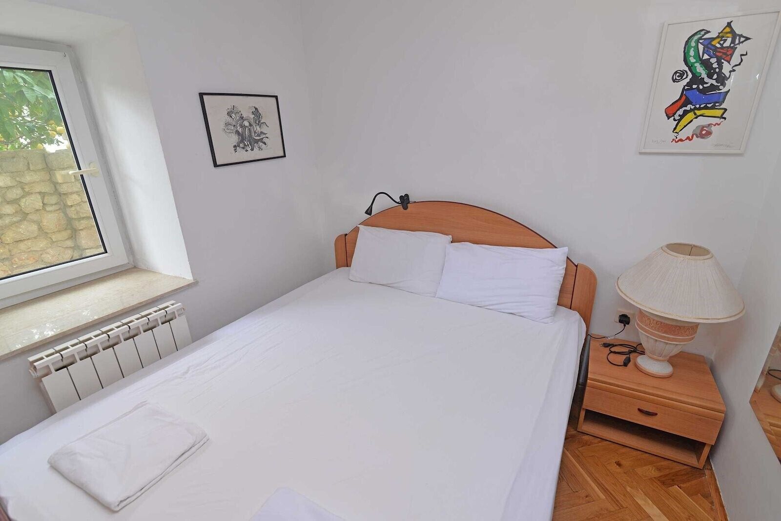 Cama bem arrumada com lençóis brancos, cabeceira de madeira, luminária de mesa e radiador sob uma janela.