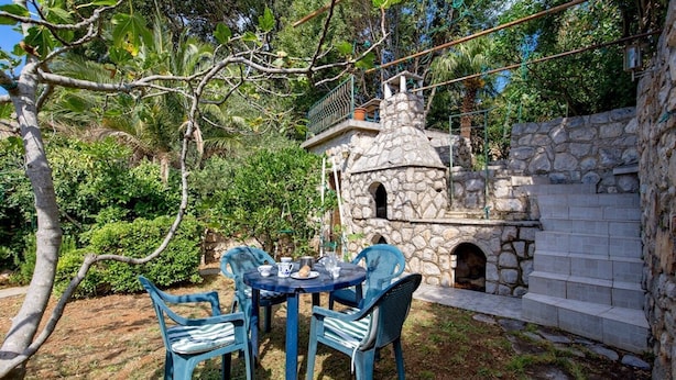 A stone-built outdoor seating area with a table and chairs, surrounded by lush greenery and a stone wall. - Studio Veli  A in Veli Lošinj (Veli Lošinj)