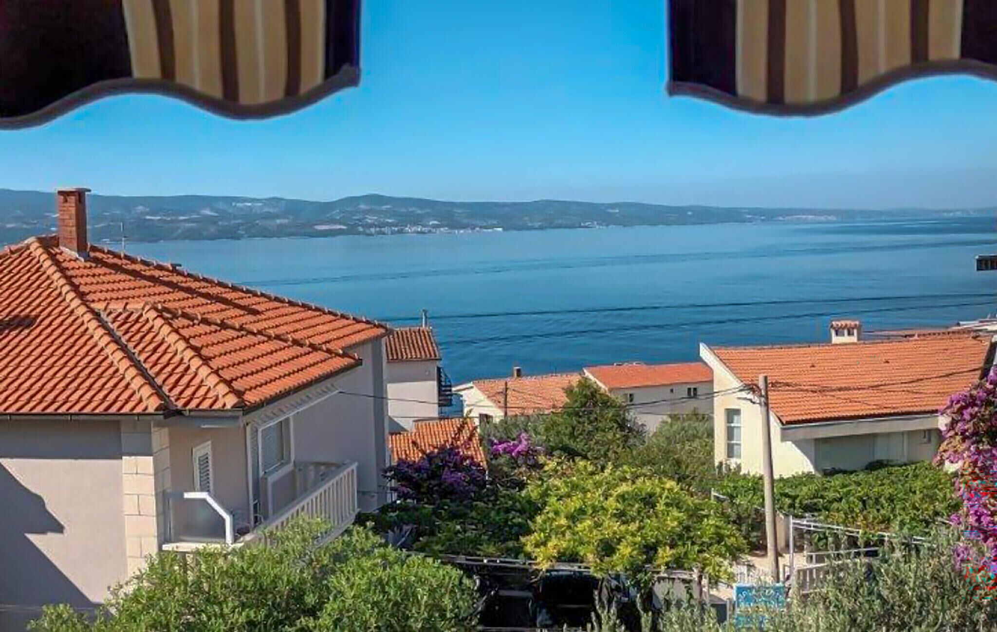 A coastal view with terracotta-roofed buildings, a clear blue sea, and distant hills.