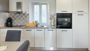Une cuisine moderne avec des meubles blancs, un four encastré et une fenêtre ornée de rideaux légers.