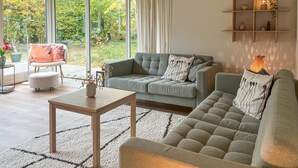 A modern living room with a grey sofa, a wooden coffee table, and a large window with curtains. - 3 bedroom gorgeous home in Sæby (Sæby)