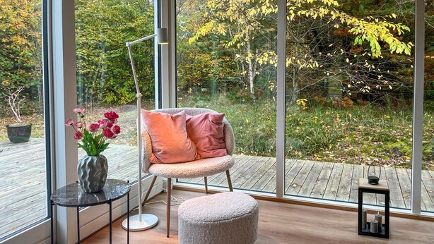 A modern room with a large window, a chair with cushions, a small round ottoman, and a table with a vase of flowers. - 3 bedroom gorgeous home in Sæby (Sæby)