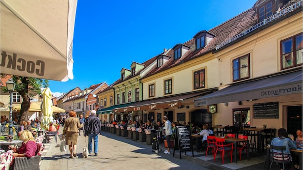 Uma rua movimentada com mesas ao ar livre em cafés e restaurantes, céu azul límpido e arquitetura europeia tradicional.