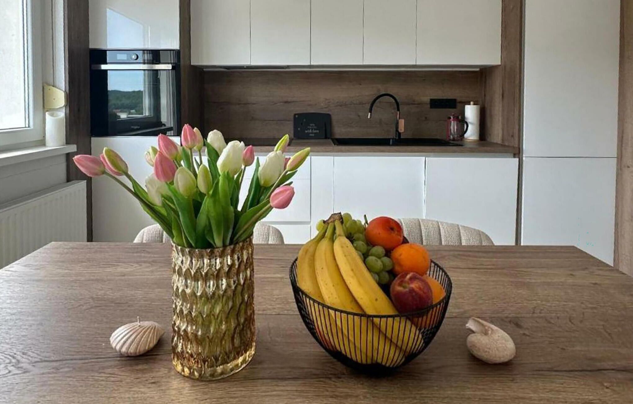 Een keuken met witte kastjes, een spoelbak en een raam. Op de tafel staat een vaas met tulpen en een schaal met fruit.