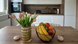 Uma cozinha com armários brancos, pia e janela. Há um vaso com tulipas e uma tigela de frutas na mesa.