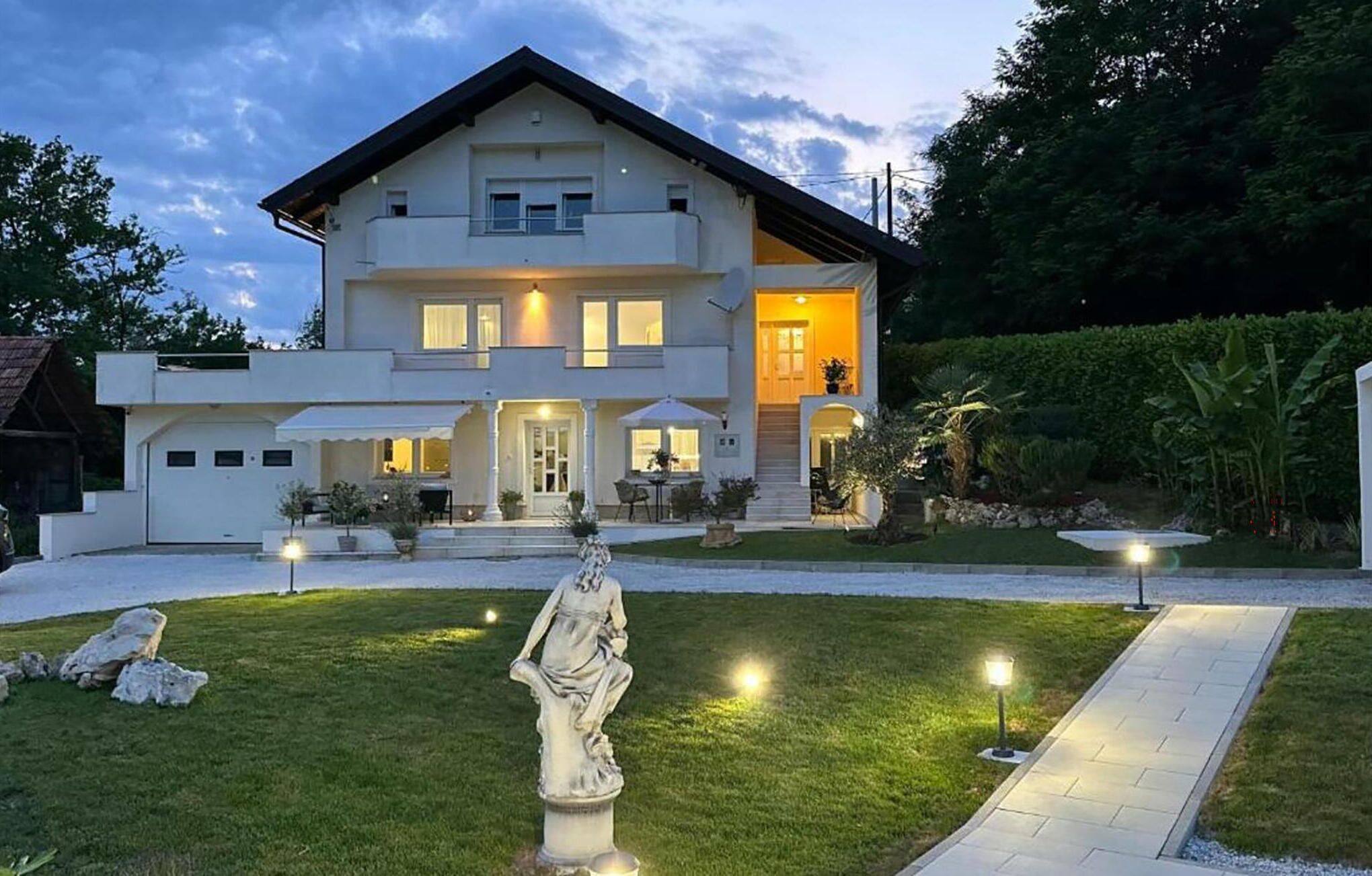 Una casa moderna de dos pisos con un césped cuidado, una estatua en la entrada y luces exteriores.