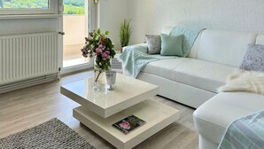Uma sala de estar moderna com um sofá branco, uma mesa de centro de vidro e vista para o verde lá fora.