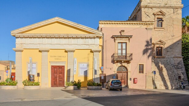 Um edifício amarelo com porta vermelha, colunas e uma placa com a inscrição "TEATRO COMUNALE NASELLI".
