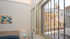 Quarto com uma cama, mesa de cabeceira com abajur, janela com vista para prédios e um quadro na parede.
