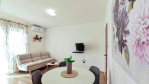 Ein Wohnzimmer mit einer Couch, einem runden Tisch mit Stühlen, einem an der Wand befestigten Fernseher und einem Blumenbild.