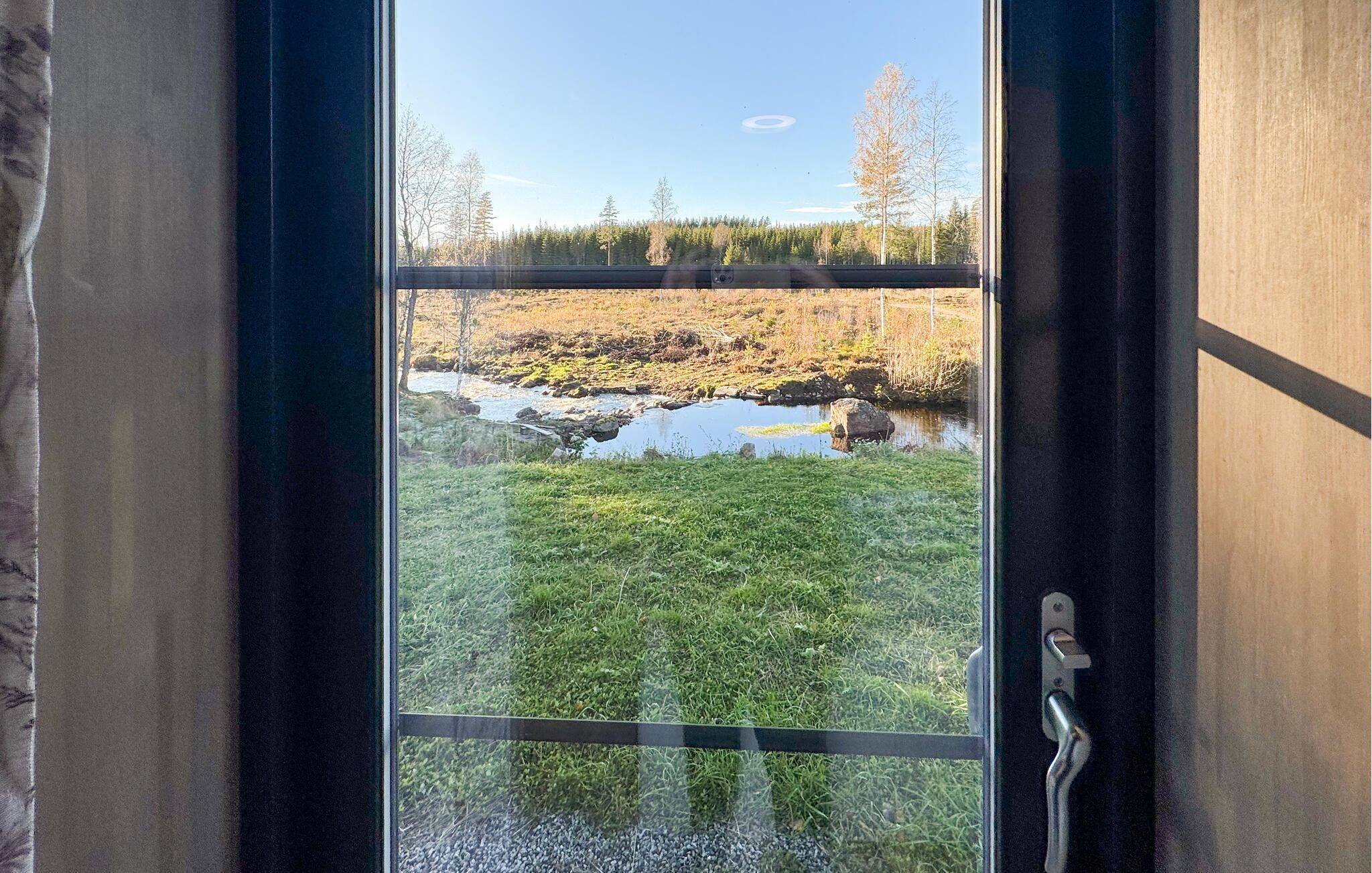 Vista a través de una ventana de metal, con un césped, un pequeño cuerpo de agua y árboles al fondo.