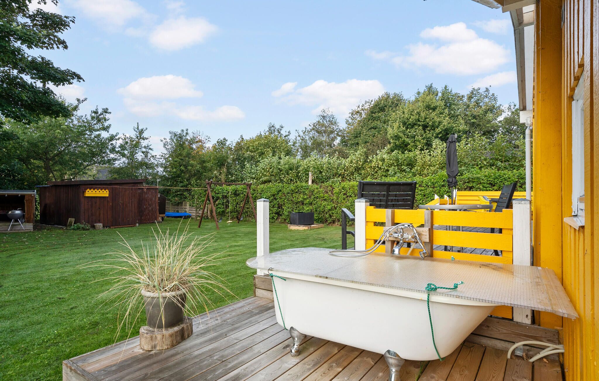 Eine Badewanne auf einer Holzterrasse mit Gartenblick, eine Holzhütte und ein gelber Zaun im Hintergrund.