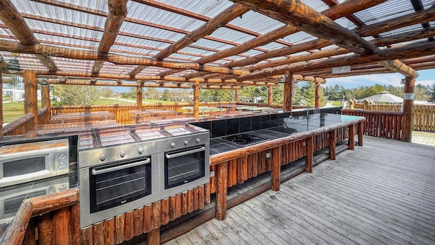 Un área de cocina exterior con terraza de madera, una parrilla metálica y un cerramiento de vidrio.