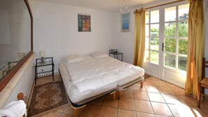 Ein Schlafzimmer mit Bett, Nachttisch, Sessel, Fenster mit Vorhängen und einem Bild an der Wand.