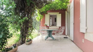 Eine Terrasse mit Tisch und Stühlen, umgeben von Pflanzen und einem Gebäude mit einem Fenster.