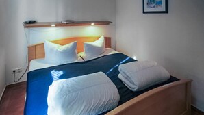Een net opgemaakt bed met witte kussens en een blauwe sprei, een houten hoofdbord en een klein plankje erboven.