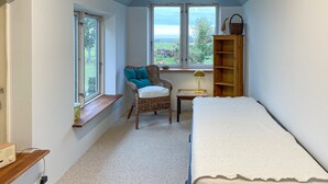 A small, cozy room with a single bed, a wicker chair with cushions, a wooden shelf, and a window with a view of greenery. - 3 bedroom gorgeous home in søby ærø (Søby Ærø)