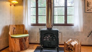 Una habitación acogedora con estufa de leña, una silla de madera con cojín, una ventana con cortinas y una lámpara fijada a la pared.