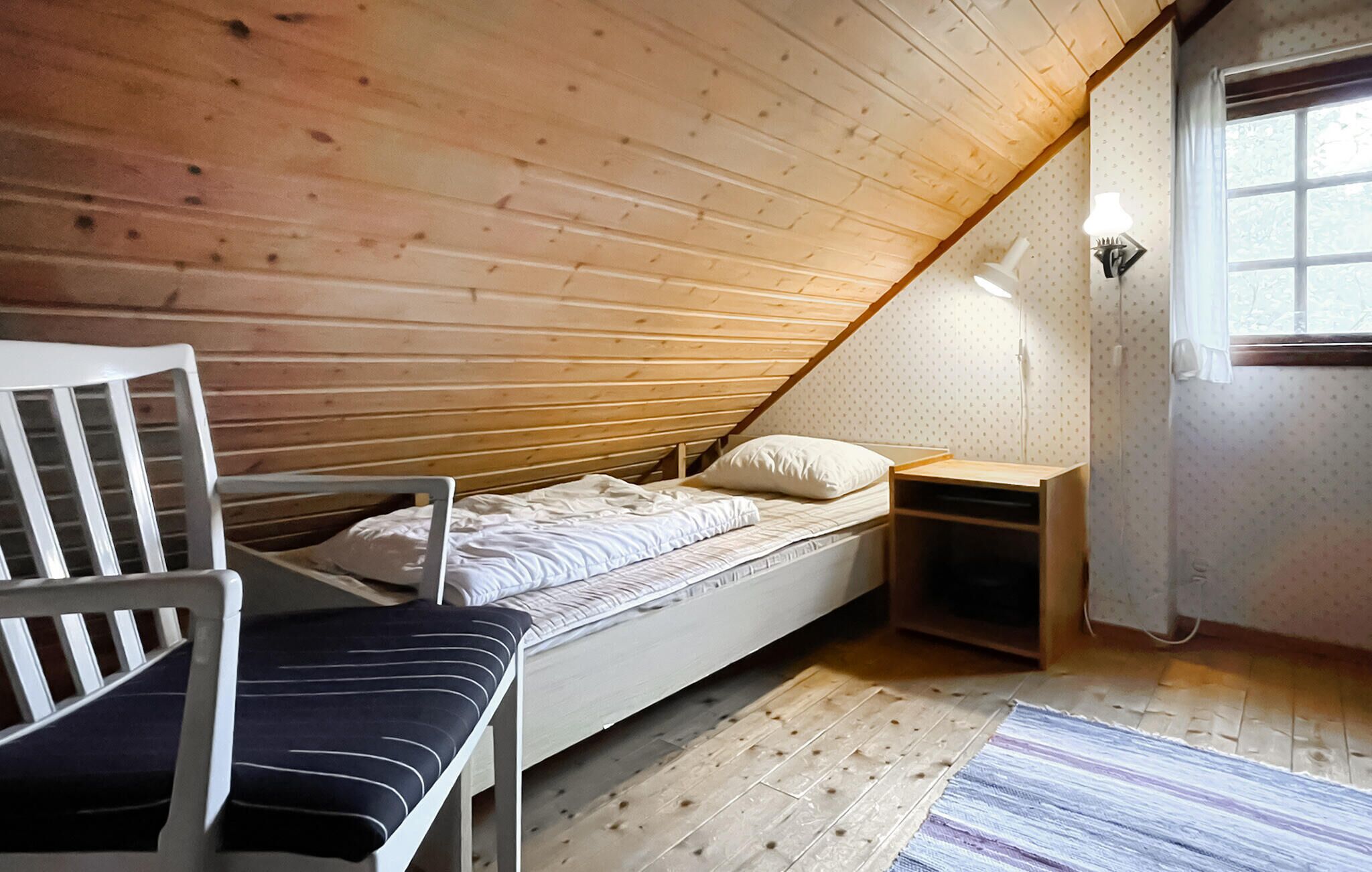 A small, sloped-roof bedroom with a single bed, a chair, a nightstand, and a window with curtains.