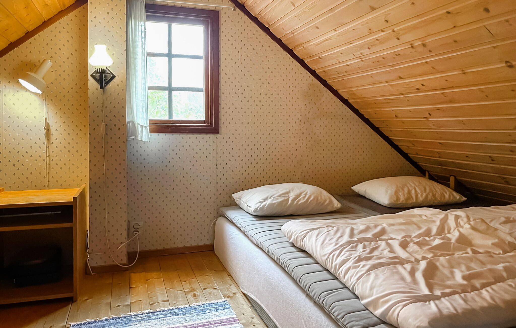Une chambre avec un plafond en pente, un lit avec une literie blanche, un bureau avec une lampe et une fenêtre avec des rideaux.