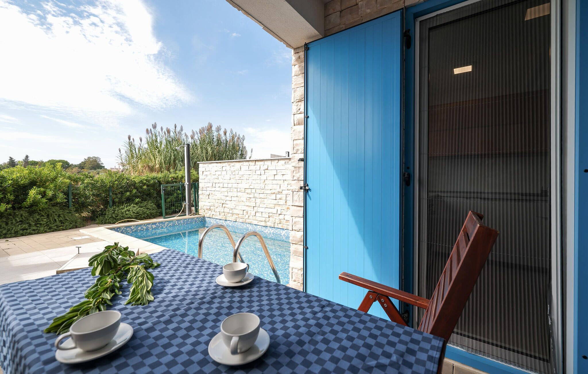 A poolside table with a checkered tablecloth, two white cups, and a green plant.