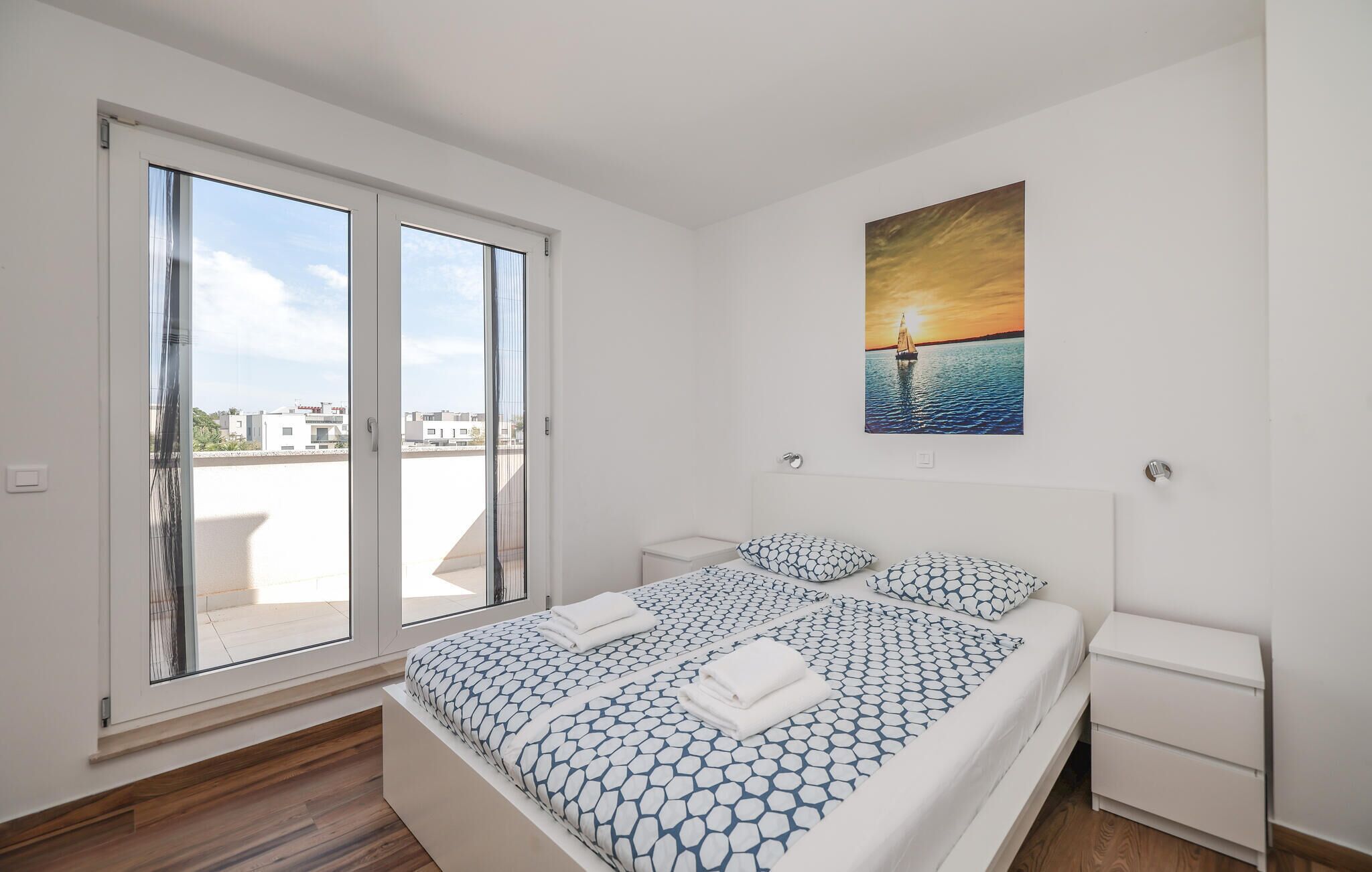 A bedroom with a large bed, two bedside tables, and a painting of a sunset over water.