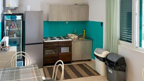 Une cuisine compacte équipée d’une cuisinière, d’un four et d’un petit évier. On y trouve également une poubelle et une fenêtre avec des stores.