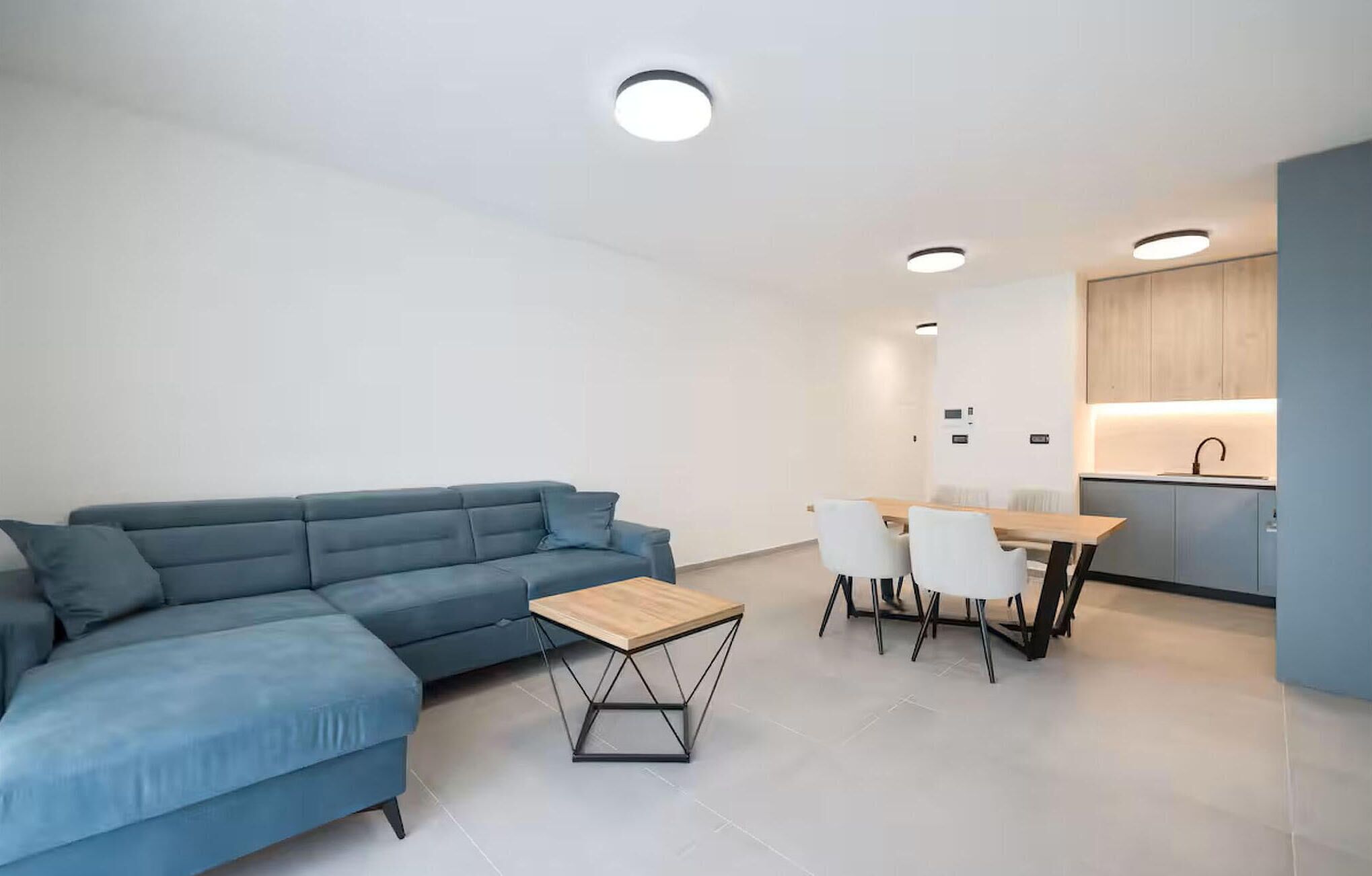 Uma sala de estar moderna com um sofá seccional azul, uma mesa de centro de madeira e uma área de jantar com cadeiras brancas e uma mesa de madeira.